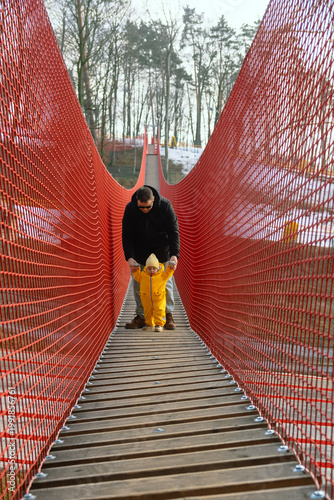 Father holding toddler on red suspension bridge in winter forest.