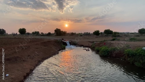 A small river winds through lush greenery, its clear water flowing gently. A lone boat drifts quietly, surrounded by trees and birdsong, creating a peaceful scene of nature’s calm beauty.