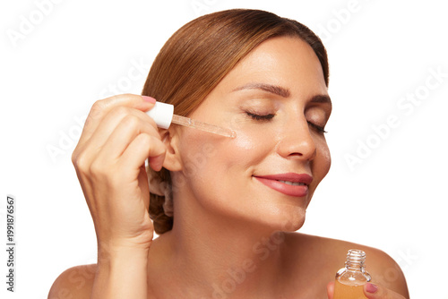 Woman smiling while gently applying skin care serum to her cheek, emphasizing a fresh beauty routine and healthy skin in a well-lit environment.