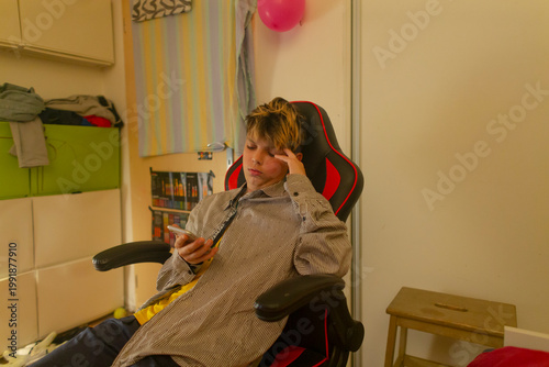 Teenager posing as rookie streamer rehearsing with phone in gaming chair, testing gestures and expressions bedroom setup hints at broadcast practice with shelf backdrop and ambient lighting