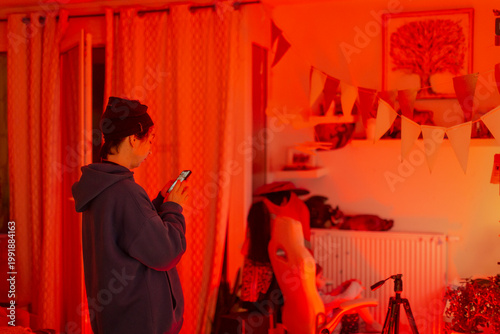 woman in warm red lit living room checking phone, surrounded by craft supplies, bunting flags, lamp glow, chair and skateboard, relaxed smile, casual hoodie, planning content