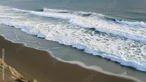 Ocean waves gently crash onto sandy beach, creating foam and ripples, with rocky cliffs visible in the background under clear blue sky