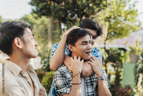 A aggressive young Asian man restraining his friend in a chokehold or rear naked choke while another watches during a tense and hostile outdoor confrontation. Outdoor shot.