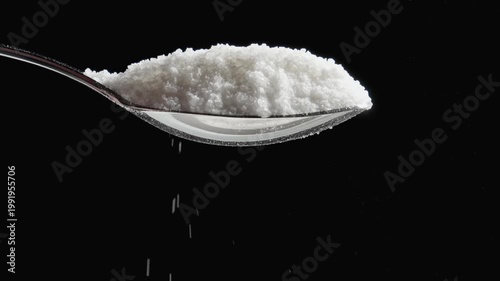 Grains of natural sugar pouring out of spoon. Sugar falling down from silver spoon black background. Spoon of white sugar. Slow motion