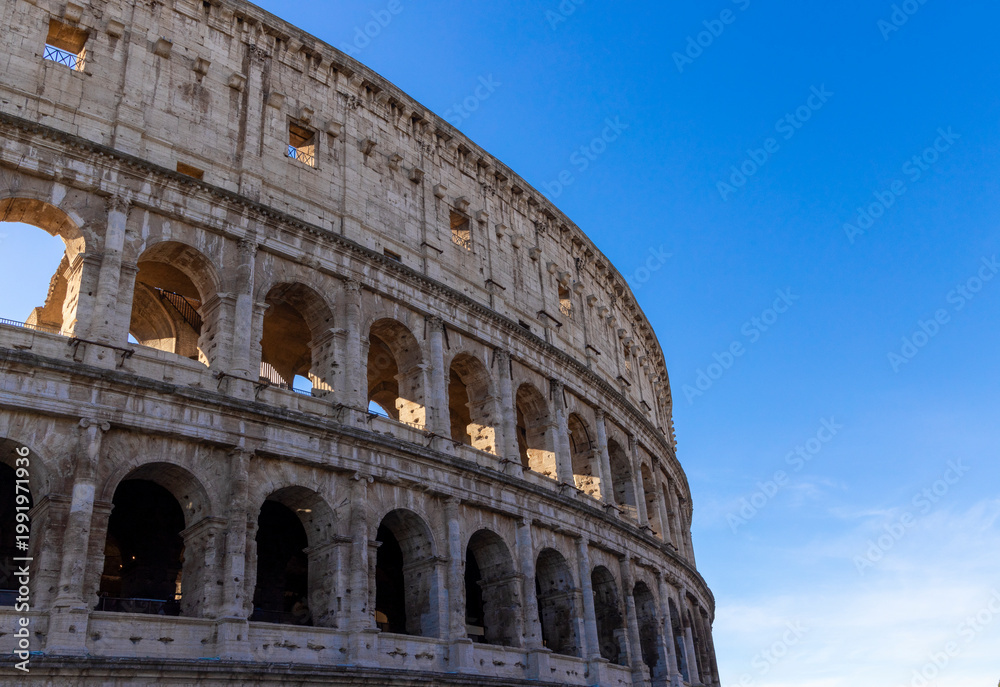 Fototapeta premium Famous Coliseum in Rome. Colosseum, or Coliseum. Part of the wall of an antique stone structure with arches. Italy, Europe. Famous touristic place in the world, Europe. Beautiful blue sky background.