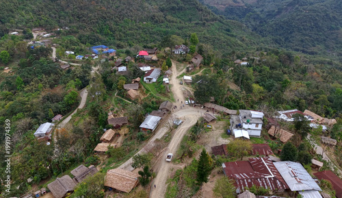 An aerial view of a rural hillside village in Nagaland, India, where winding dirt roads connect scattered thatched and tin-roofed houses across forested slopes, reflecting remote living