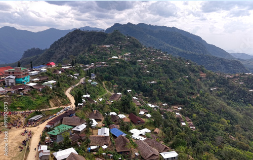 An aerial view of a hillside village in Nagaland, India, with thatched and tin-roofed houses spread along a winding road, as people gather along the roadside, set against forested mountain layers