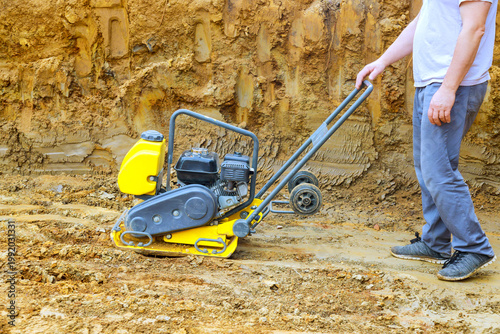 Fotografía Construction worker pushes plate compactor over loose soil at building site in dust rises as machine flattens ground for paving