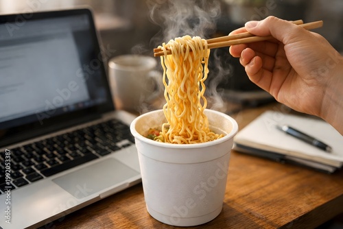 Steam rises from a cup of noodles as someone uses chopsticks to lift the noodles. A laptop sits open next to a notebook and a cup in a busy workspace