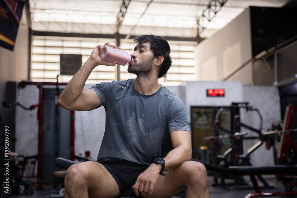 Naklejka premium Tired young man drink water from bottle to rest after heavy workout in gym. Athlete covered in sweat look exhausted but stay determined to achieve strong fitness health goal