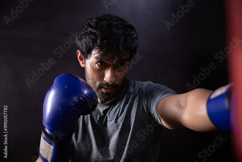 Determined male boxer training hard punching heavy red bag wearing blue boxing glove looking focused and aggressive with sweaty face during intense workout session in dark gym environment