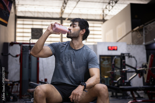 Tired young man drink water from bottle to rest after heavy workout in gym. Athlete covered in sweat look exhausted but stay determined to achieve strong fitness health goal