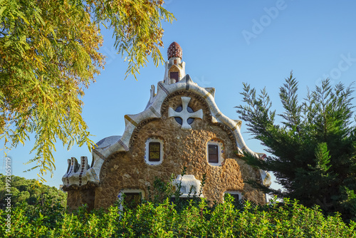 Fotografie Park Guell,Barcelona,Spain,A unique stone building with organic architectural design, featuring a cross emblem and surrounded by greenery under a clear blue sky