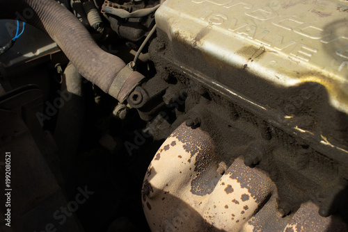 A detailed, macro-style photograph of a vehicle's engine bay, showing various dusty components including wires, hoses, and mechanical parts