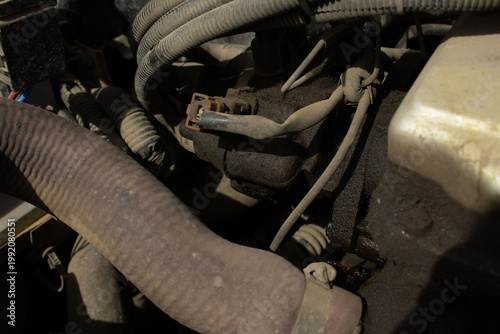 A detailed, macro-style photograph of a vehicle's engine bay, showing various dusty components including wires, hoses, and mechanical parts