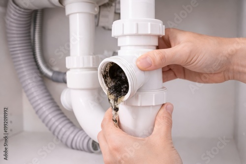 Clearing Clogged Sink Drain Trap At Home. Hands open a sink trap to remove a blockage and dirty water during routine household plumbing maintenance under the basin.