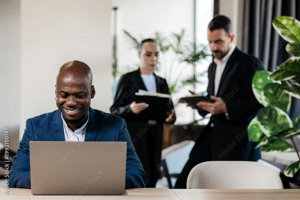 Obraz premium African American man in suit working at desk with laptop, colleagues chatting in background. Office workflow concept.