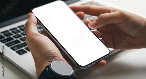 Hands holding smartphone with blank screen over laptop keyboard, copy space