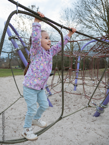 Young smiling girl in purple fleece jacket holds the thick ropes of a climbing net at outdoor playground. Vertical orientation
