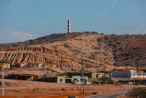 A wide-angle landscape photograph featuring a distinctive striped lighthouse perched atop a sun-drenched, arid hill. The foreground shows a rural coastal village with rugged terrain, sparse vegetation