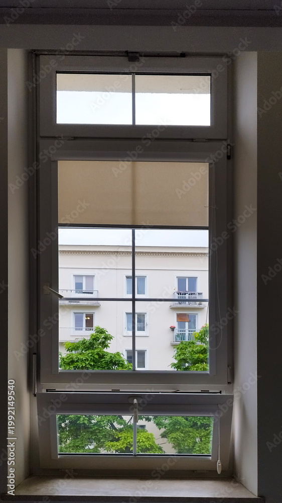 Fototapeta premium A window shows an outside view of a building and trees on a cloudy day in a city
