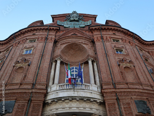 Foto Prospettiva dal basso della facciata barocca in mattoni di Palazzo Carignano con il balcone monumentale e le bandiere istituzionali sotto un cielo azzurro, Torino, Italia