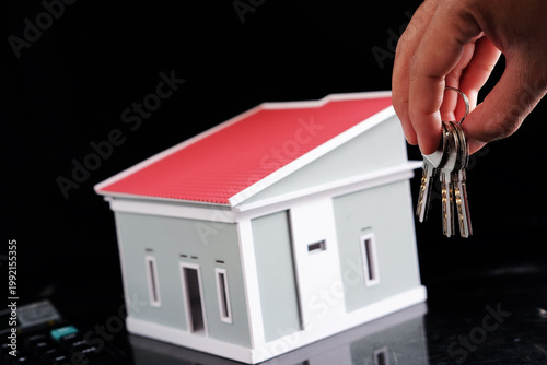 Close-up of a person holding a house key next to a miniature house. Concept of handover, purchase, and rental of housing.
