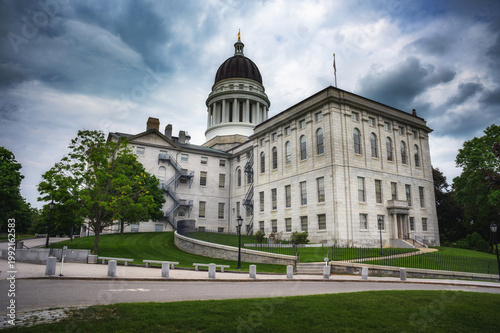 Side perspective of the historic Maine State Capitol building in Augusta, showing the dome, stone facade, windows, and landscaped grounds under dramatic cloudy sky.