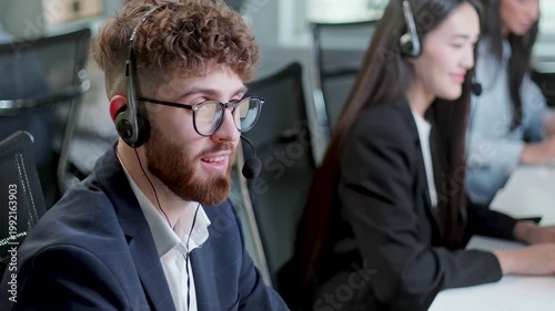 Young male customer support agent speaking through headset while working at computer in a modern call center office. Professional help desk employee communicates with clients, while colleagues in the