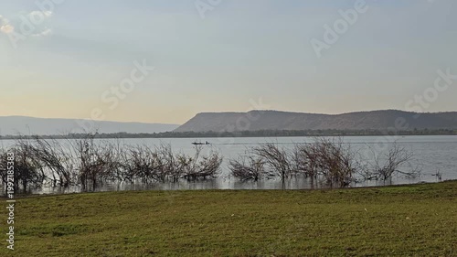 Calm river boat by lake shoreline with grass, distant hill and flooded shrubs under soft hazy sky