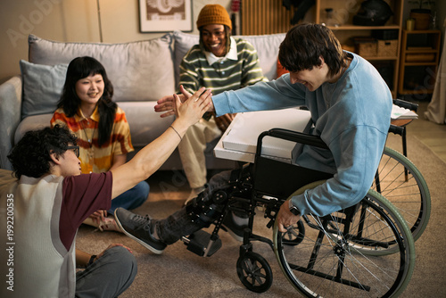 Εκτύπωση καμβά Young adult man with disability in wheelchair high fiving male friend while diverse friends gathering at home, representing inclusion and support
