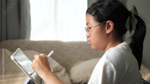 Profile view of a young Asian girl with glasses and a cute hair clip, focused on using a tablet and stylus while relaxing on a sofa at home, e-learning and digital lifestyle concept