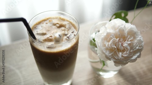 Close-up of refreshing iced latte coffee with a black straw and a blooming white rose in a glass vase on a wooden table, aesthetic cozy cafe lifestyle concept