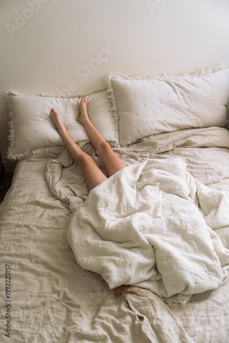 Young girl lying in bed under crumpled linen bedding with legs playfully resting on pillows, cozy minimal morning aesthetic. Bed rotting