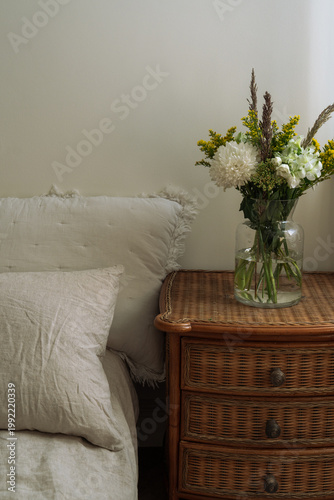 Cozy bedroom scene with oatmeal linen bedding, pillows and a glass vase of fresh flowers on a wicker nightstand, soft natural light.