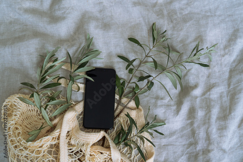 Flat lay of mesh eco bag with smartphone and olive tree branches on oatmeal linen bedding, soft light and earthy colours creating a minimal aesthetic composition.