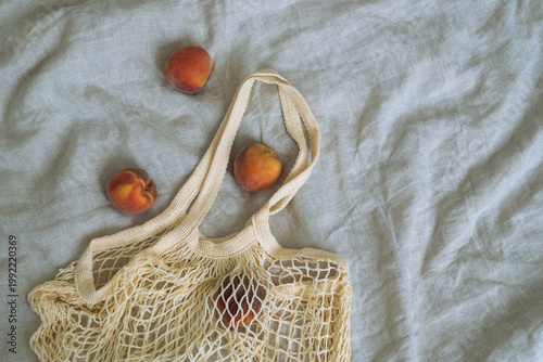 Flat lay of mesh eco bag with a few fresh peaches on oatmeal linen bedding, soft light and earthy colours creating a minimal aesthetic composition with copy space