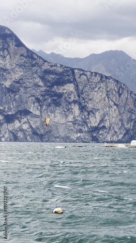 Kitesurfer riding on windy Lake Garda with strong waves in stormy weather. Small island in the background and distant Alps under dramatic cloudy sky.