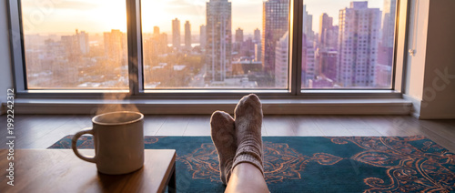 Feet resting on a rug by the window while drinking hot coffee and watching city sunset