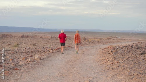 older European couple walk away from camera, along rocky desert road into vast Namibian landscape