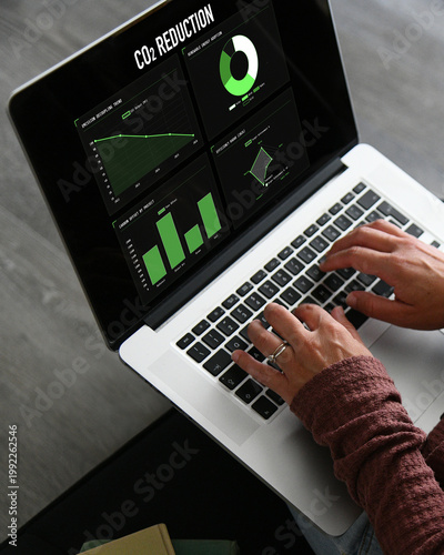 A professional woman uses her laptop to analyze a CO2 reduction dashboard, monitoring environmental data and carbon footprint metrics through various digital charts and green technology graphs online.