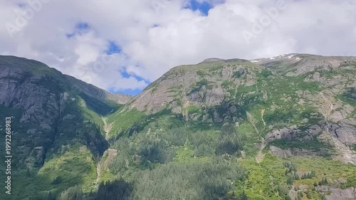 August 13, 2024 - Tracy Arm, USA - A view of mountains covered with forests and a valley with a small stream flowing through. Some clouds dot the sky. Mountains are partially covered in snow.