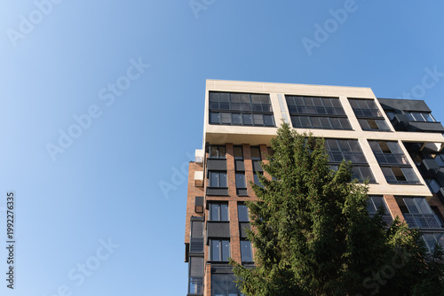 Low angle view of a modern residential building with brick and light facades, framed by an evergreen tree against a clear blue sky with ample copy space.