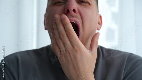 Close-up of a young man yawning in a bright room, showing signs of sleepiness in the morning. Concept of fatigue and waking up.