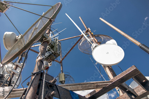 A complex array of telecommunication antennas and satellite dishes mounted on a metal structure. The low-angle shot highlights industrial connectivity against a clear blue sky.