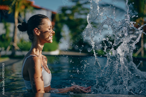 Fotografi A smiling woman enjoys a refreshing moment at an outdoor pool, water spraying as a fountain arcs nearby