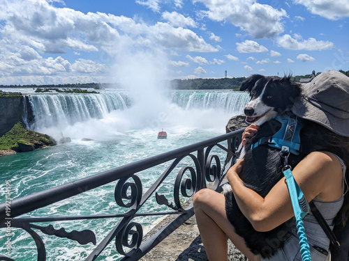 A dog looks at the camera while being held by a woman overlooking Niagara Falls with a maid of the mist boat on the river during a sunny summer day.