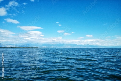 Wide panoramic view of Lake Skadar surface with blue rippled water and distant mountains on the horizon under a clear blue sky with white clouds in Montenegro, Southeastern Europe region