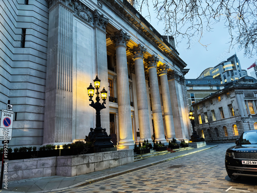 Naklejka premium Grand neoclassical Four Seasons Hotel London at Ten Trinity Square building with illuminated Corinthian columns and ornate lamp posts in London, UK evening cityscape