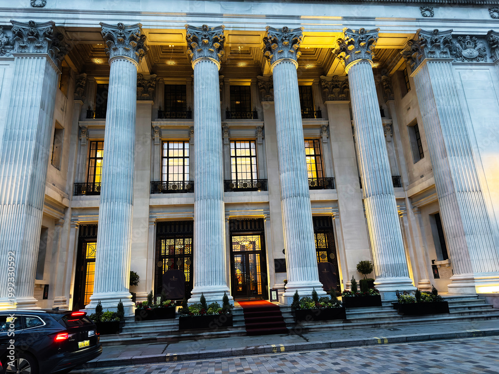 Naklejka premium Grand neoclassical building with Corinthian columns and illuminated hotel entrance in London UK at dusk on a city street. Four Seasons Hotel London at Ten Trinity Square.